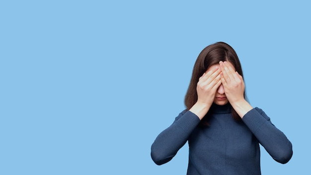 A frustrated female student in glasses and a sweater covering her eyes with her hands on a blue background