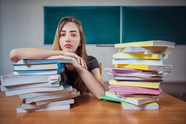 Frustrated female student sitting at the desk with a huge pile of study books in classroom