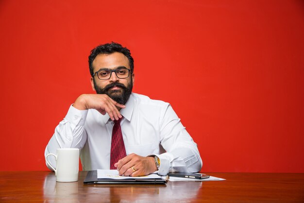 Разочарованный индийский или азиатский молодой бизнесмен с бородой, представляя что-то, сидя за офисным столом