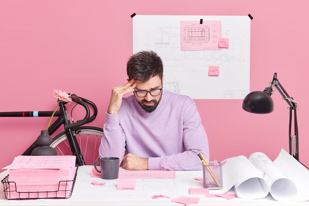 Frustrated male architect works on sketch focused at papers creats sketches for future building project poses in coworking space dressed in casual clothes