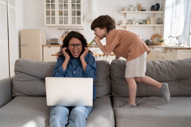 Frustrated mother of hyperactive child scream of annoying chaos\
work on laptop with disobedient son