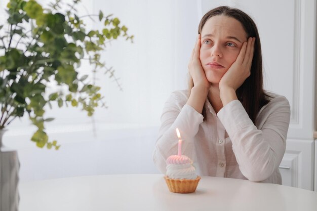 イライラした悲しい白人の茶色の髪の女性が家の内部で誕生日ケーキを持ってテーブルに座って、休日を一人で祝うために動揺している不幸な表情で目をそらしている