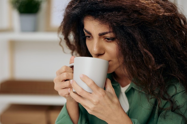 Frustrated sad tanned curly latin lady troubled with problem\
with cup of tea coffee in casual shirt sit on chair in home modern\
interior background look aside copy space banner concept\
psychology