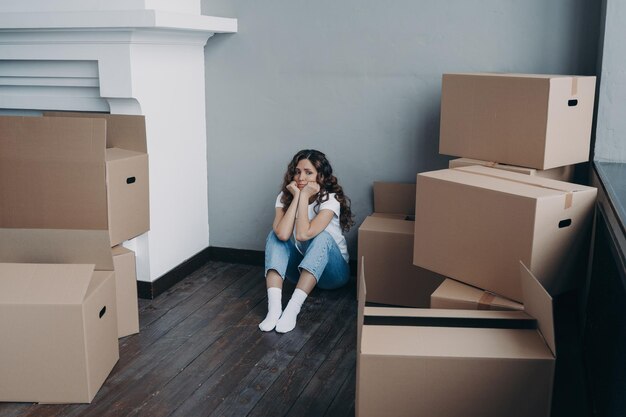 Frustrated woman is looking at packed boxes sadly and sitting\
on floor girl is upset with moving