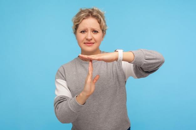 Frustrated woman with short curly hair in grey sweatshirt feeling tired and overworked showing timeout gesture need more time missing deadline indoor studio shot isolated on blue background