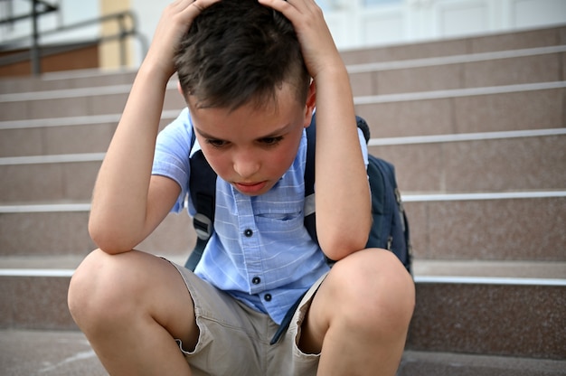 Scolaro preoccupato frustrato che si tiene la testa con le mani mentre è seduto sulle scale vicino alla scuola. concetto di difficoltà di apprendimento, bullismo, solitudine