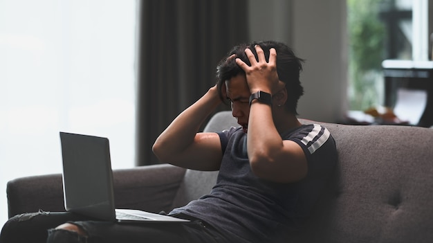 Frustrated young Asian man sitting on sofa with laptop and feeling stress from work