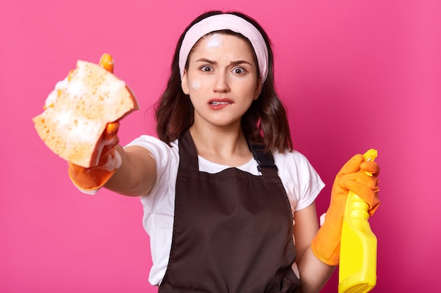 Frustrated young brunette female holds sponge and detergent, biting her lip, feeling stressed