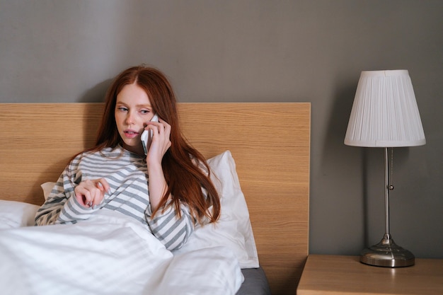 Frustrated young business woman sitting on cosy bed at home and talking mobile phone before bedtime in evening at home