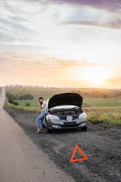 A frustrated young girl stands near a broken-down car in the\
middle of the highway during sunset. breakdown and repair of the\
car. waiting for help.