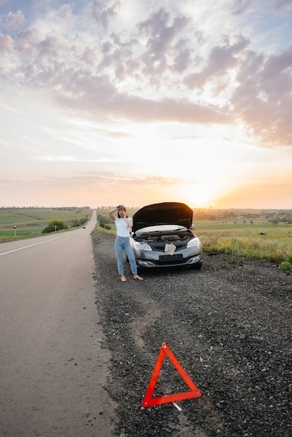 A frustrated young girl stands near a broken-down car in the\
middle of the highway during sunset. breakdown and repair of the\
car. waiting for help.