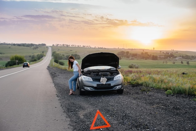 A frustrated young girl stands near a broken-down car in the middle of the highway during sunset. Breakdown and repair of the car. Waiting for help.