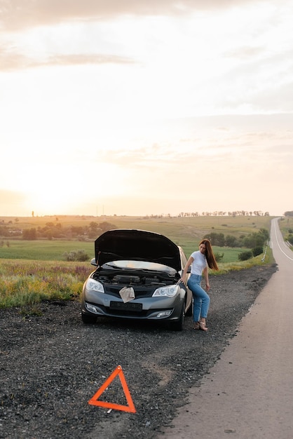 A frustrated young girl stands near a brokendown car in the\
middle of the highway during sunset breakdown and repair of the car\
waiting for help car service car breakdown on road