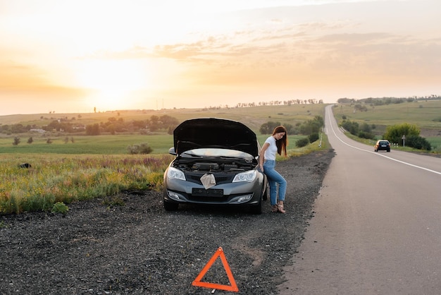 A frustrated young girl stands near a brokendown car in the middle of the highway during sunset Breakdown and repair of the car Waiting for help Car service Car breakdown on road