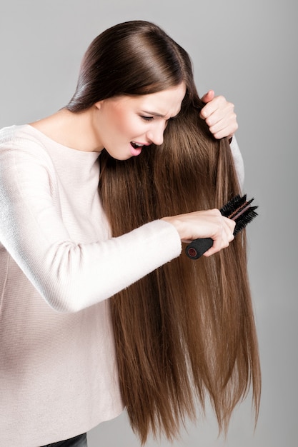 Frustrated young woman combing tangled long natural hair with hairbrush