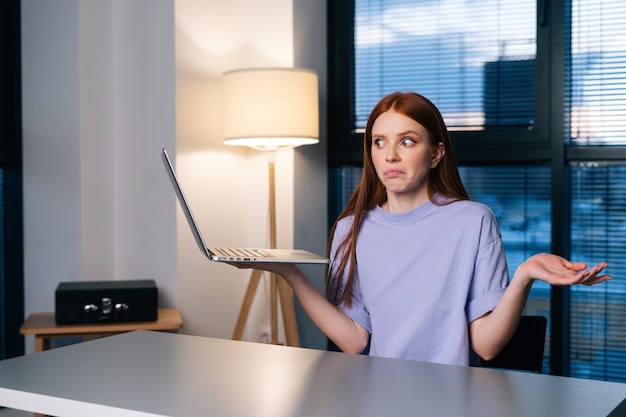 Frustrated young woman confused by laptop computer problem sitting at desk in home office near window evening at late.