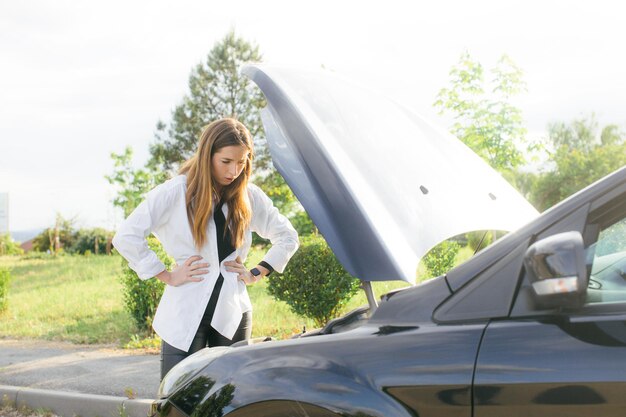 Frustrated young woman looking at broken down car engine on street