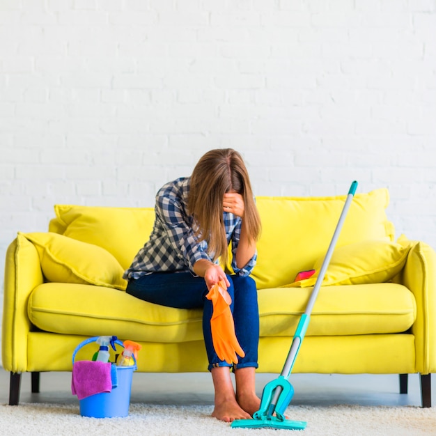 Frustrated young woman sitting on sofa with cleaning equipments