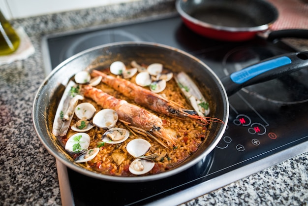 Foto padella su un piano cottura in vetroceramica con paella fatta in casa appena fatta a casa. riso con gamberi e vongole.