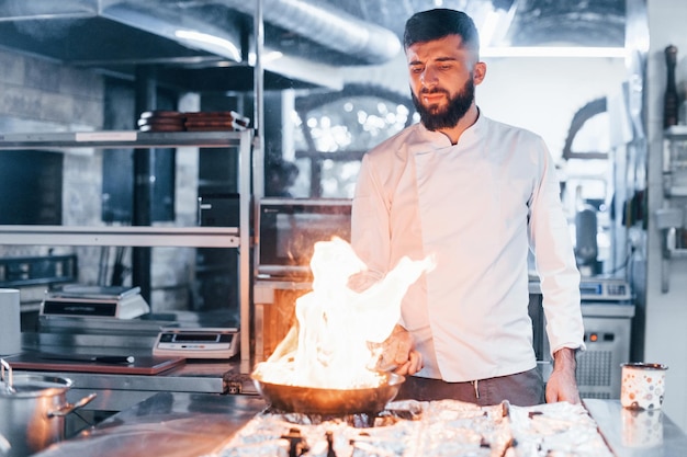 Frying pan is on fire Chef in white uniform cooking food at kitchen Busy day at work