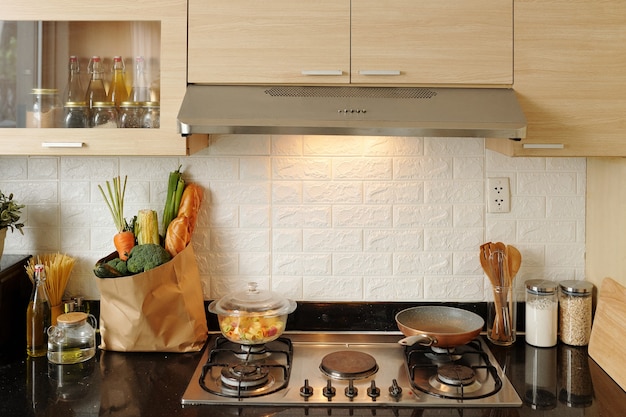 Frying pan and saucepan with pasta on kitchen stove and bag of fresh groceries on counter