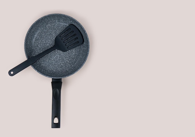 Photo frying pan with black nonstick coating and black polished kitchen spatula on beige background