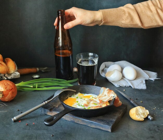 Una padella con uova strapazzate e pancetta sul tavolo della cucina a casa e una birra in mano