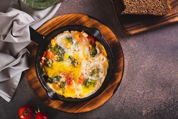 Frying pan with scrambled eggs with broccoli and tomatoes on a wooden board top view