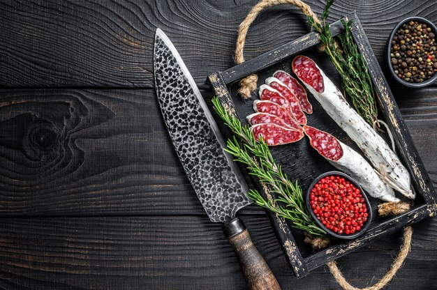 Fuet Salami sausage slices with thyme and rosemary in a wooden tray. Black wooden background. Top view. Copy space.