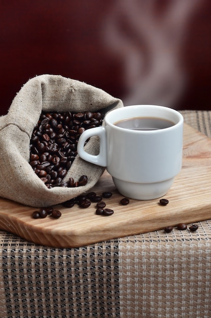 На деревянной поверхности лежит полный пакет коричневых кофейных зерен и белая чашка горячего кофе.