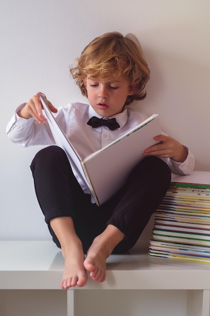 Full body of adorable barefoot boy in formal clothes reading interesting book while sitting on white background in light room