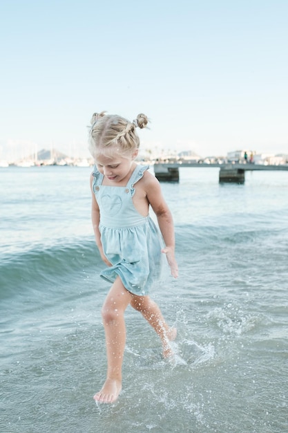 Full body of adorable barefoot girl entertaining in clean wavy water of sea on shore on sunny day in summer