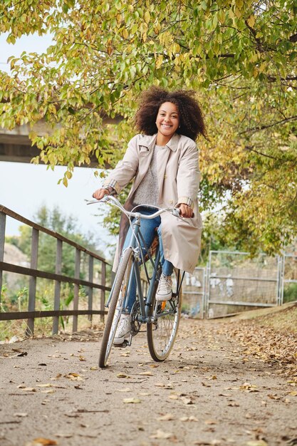 秋の日中に道で自転車に乗るカジュアルな服を着た巻き毛のアフリカ系アメリカ人女性の全身