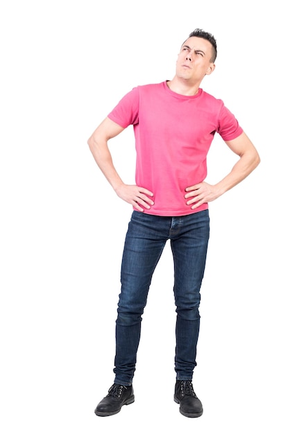Full body of attentive young male model with dark hair in casual clothes standing against background with hands on waist and looking away with hesitation