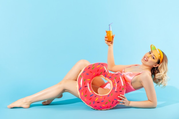 全身、魅力的な水着姿の幸せな女性がセクシーに横たわり、カメラに微笑んで、ゴムの指輪、フレッシュジュースまたはカクテルを持って、夏のビーチ、休暇リゾートでリラックス。屋内スタジオショット分離
