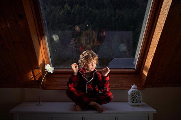 Full body of barefoot cute preteen boy meditating in Padmasana yoga pose while sitting with crossed legs on chest of drawers in evening