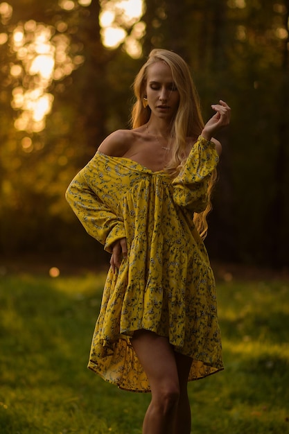 Full body blond female in floral dress looking looking away while standing on grass in evening in forest