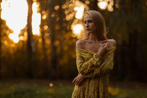 Full body blond female in floral dress looking looking away while standing on grass in evening in forest