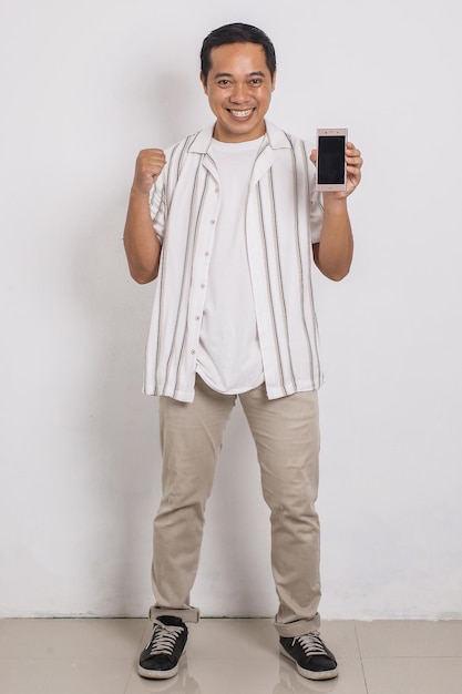 full body of casual asian man in shirt showing his smartphone blank screen with winning gesture