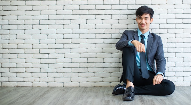 Full body cheerful Asian male manager smiling and looking at camera while relaxing near gray brick wall