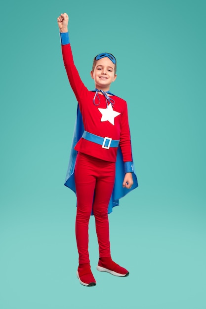 Photo full body cheerful boy in superhero costume raising clenched fist and smiling for camera while being ready to save world against turquoise