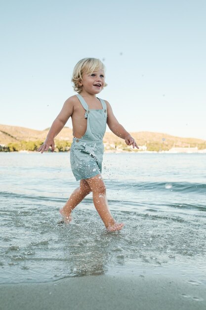 Full body of cheerful cute little child in summer clothes walking and looking away while smiling in calm water of warm sea