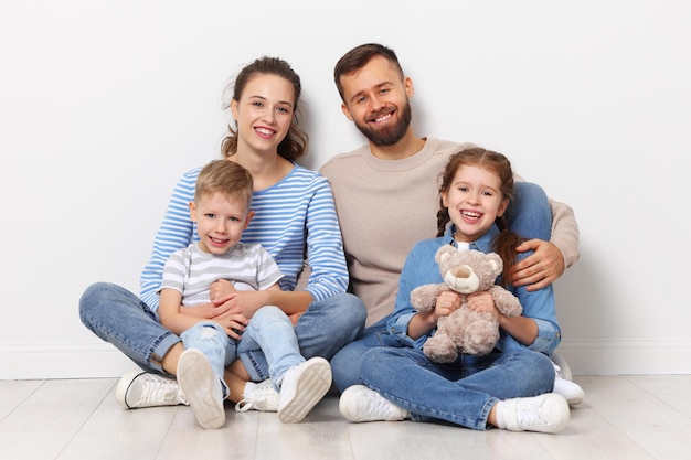 Full body of cheerful family with little kids looking at camera\
while sitting together on floor near wall in empty room of new\
home