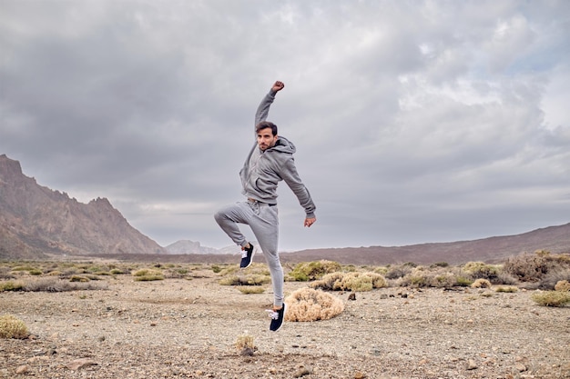 Full body of cheerful male in sportswear jumping on dry ground near volcanic area of mountain Teide in Tenerife Canary Islands in Spain in cloudy day