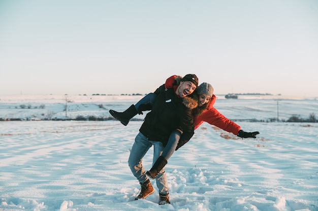 Full body of cheerful man giving piggyback ride and falling and laughing with girlfriend in snowy countryside