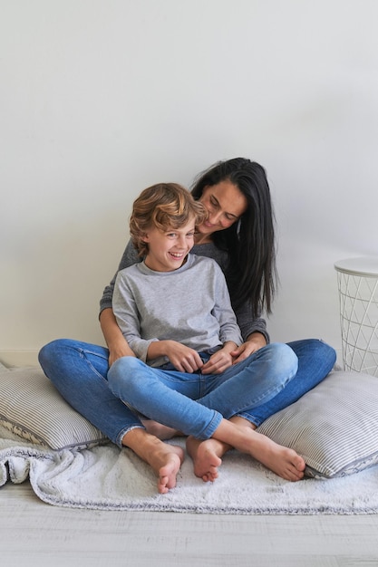 Full body of cheerful preteen boy and dark haired woman hugging and laughing while resting on floor in living room