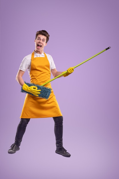 Full body of cheerful young male in yellow apron and latex gloves pretending playing music on mop during household chores against violet background