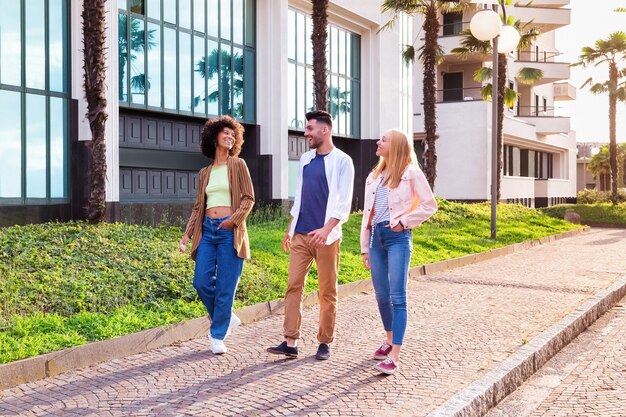 Full body of cheerful young multiracial students in trendy casual outfits chatting and laughing friendly while strolling together on pavement near modern buildings in summer day in city