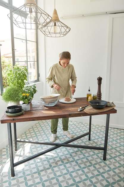 Full body of concentrated female cook in uniform putting steak tartare on white serving plate in light kitchen