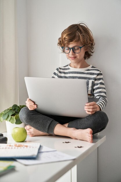 Полное тело сосредоточенного ребенка-подростка, читающего информацию на нетбуке и смотрящего на экран во время онлайн-обучения в спальне